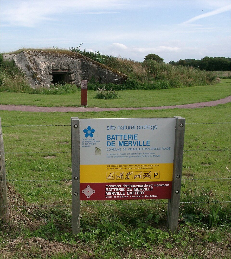 Casemate en béton de la Batterie de Merville, enterrée sous un talus herbeux, avec le panneau du site classé au premier plan.