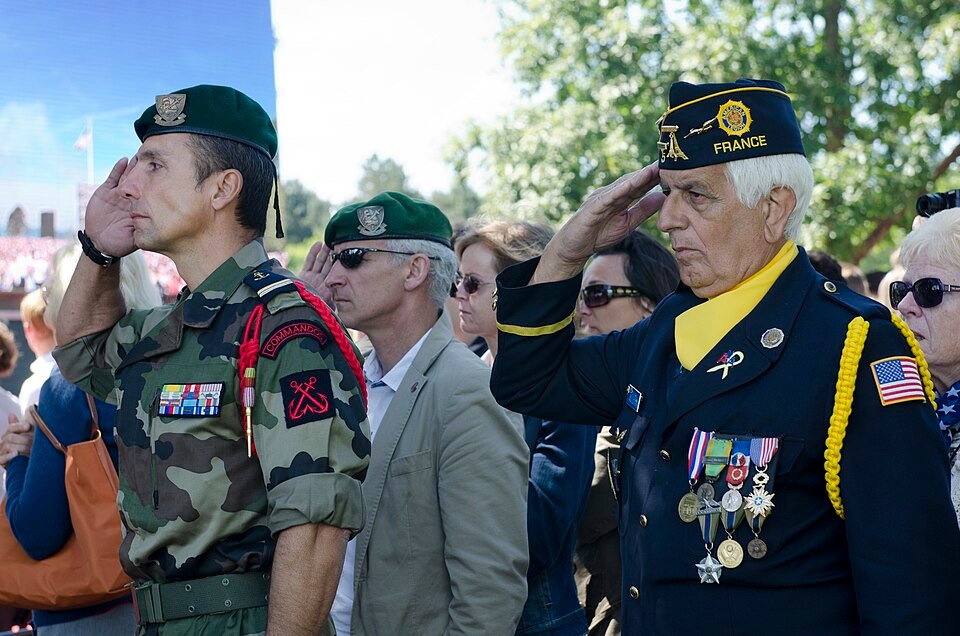 Un militaire français en béret vert et un vétéran américain décoré saluent côte à côte lors d'une cérémonie du 6 juin en Normandie.