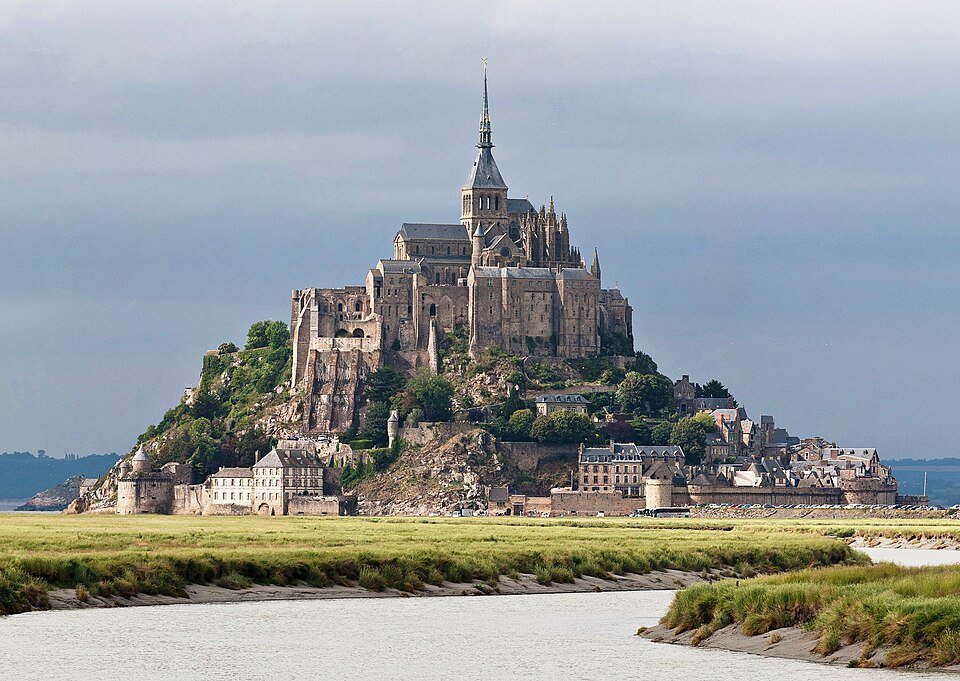 Le Mont-Saint-Michel et son abbaye vus depuis les herbus de la baie, à marée basse, sous un ciel couvert.
